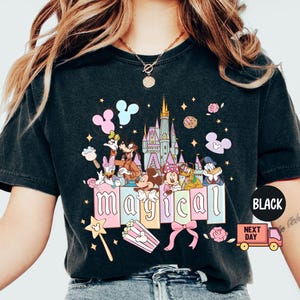 May include: Black t-shirt featuring a colorful graphic of Disney characters, a castle, and the word "magical". The design includes Mickey and Minnie Mouse, Donald Duck, and Goofy. A pink bow, popcorn, and a magic wand are also included.