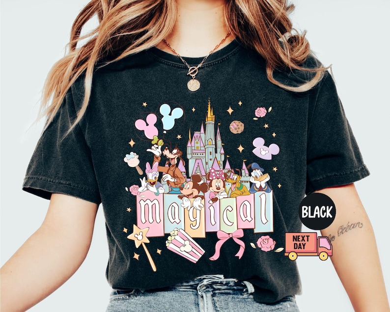 May include: Black t-shirt featuring a colorful graphic of Disney characters, a castle, and the word "magical". The design includes Mickey and Minnie Mouse, Donald Duck, and Goofy. A pink bow, popcorn, and a magic wand are also included.