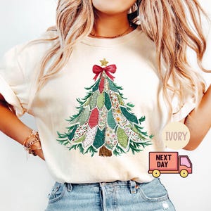 May include: Ivory-colored t-shirt featuring a Christmas tree design. The tree is composed of various fabric patches in green, red, and floral patterns, topped with a red bow and gold star. The shirt has short sleeves and a relaxed fit.
