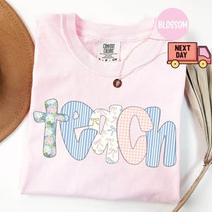 May include: A light pink Comfort Colors t-shirt with the word "TEACH" appliqued across the front. The letters are made of various patterned fabrics, including floral, gingham, and stripes. A gold necklace with a pendant is visible.