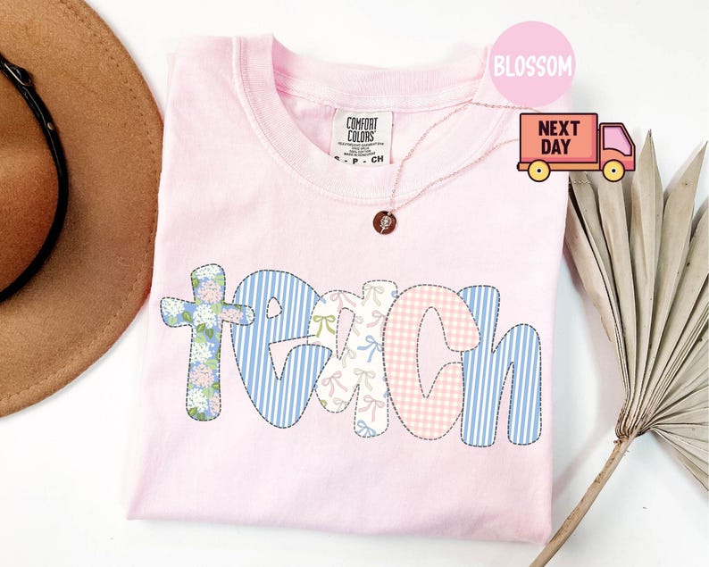 May include: A light pink Comfort Colors t-shirt with the word "TEACH" appliqued across the front. The letters are made of various patterned fabrics, including floral, gingham, and stripes. A gold necklace with a pendant is visible.