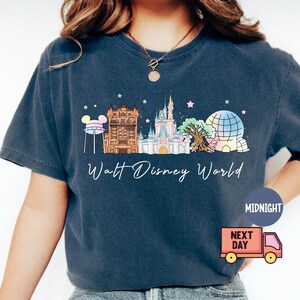 May include: Navy blue t-shirt featuring a colorful illustration of Walt Disney World landmarks, including Cinderella Castle and Spaceship Earth. The text "Walt Disney World" is written in a script font. The shirt has a relaxed fit.