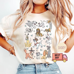 May include: Ivory-colored t-shirt featuring a collection of cartoon dog illustrations. The dogs are in various poses and styles, with some wearing hats. The shirt has a round neckline and short sleeves. The text "IVORY" is printed on the shirt.