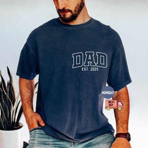 May include: Navy blue t-shirt with the word "DAD" in white block letters and "EST. 2025" below it. The shirt is a relaxed fit and has short sleeves. A small graphic is on the lower right side.