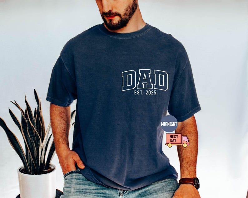May include: Navy blue t-shirt with the word "DAD" in white block letters and "EST. 2025" below it. The shirt is a relaxed fit and has short sleeves. A small graphic is on the lower right side.