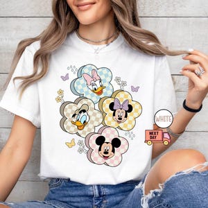 May include: White t-shirt featuring a floral design with cartoon characters. The design includes Mickey Mouse, Minnie Mouse, Donald Duck, and Daisy Duck, each within a flower-shaped frame with a checkered pattern. Butterflies and flowers are also included.