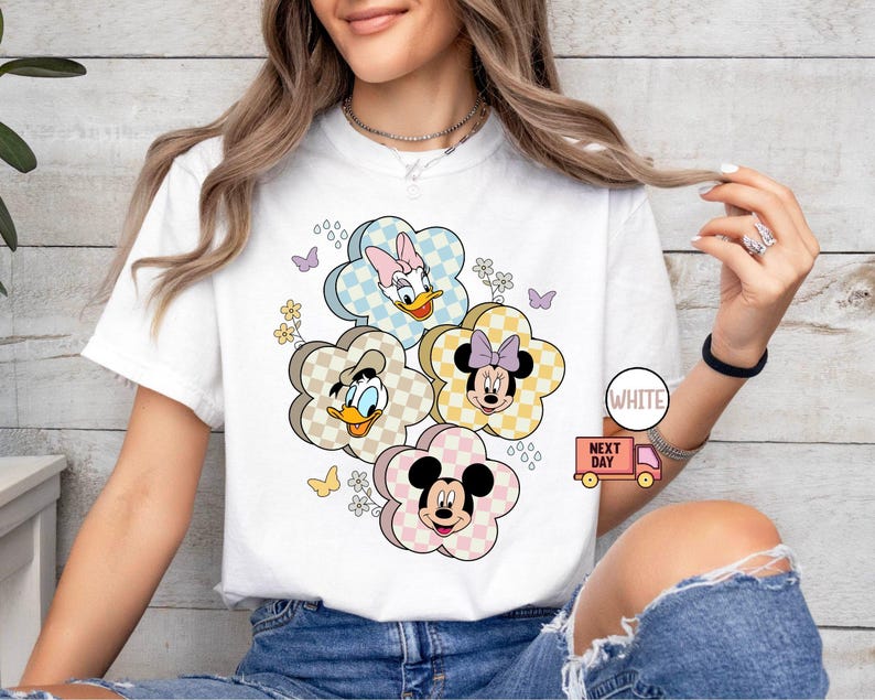 May include: White t-shirt featuring a floral design with cartoon characters. The design includes Mickey Mouse, Minnie Mouse, Donald Duck, and Daisy Duck, each within a flower-shaped frame with a checkered pattern. Butterflies and flowers are also included.