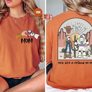 Pu&ograve; includere: Una t-shirt arancione con una grafica frontale di due mani che formano un cuore con il testo MOM sotto. La grafica sul retro raffigura due personaggi dei cartoni animati in un paesaggio desertico con il testo YOU GOT A FRIEND IN ME.