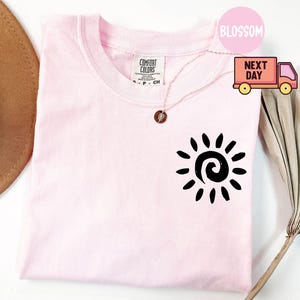 May include: A light pink Comfort Colors t-shirt with a black sun graphic on the front. The shirt has a crew neck and short sleeves. A gold necklace with a pendant is draped across the shirt. The word "Blossom" is in a pink circle.