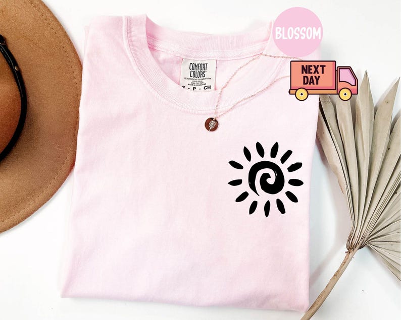 May include: A light pink Comfort Colors t-shirt with a black sun graphic on the front. The shirt has a crew neck and short sleeves. A gold necklace with a pendant is draped across the shirt. The word "Blossom" is in a pink circle.