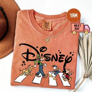 K&ouml;nnte beinhalten: Korallfarbenes T-Shirt mit dem Wort "Disney" in Schwarz, mit Zeichentrickfiguren, die einen Zebrastreifen &uuml;berqueren. Zu den Figuren geh&ouml;ren Pluto, Goofy, Donald Duck und Mickey Mouse. Eine goldene Halskette und ein brauner Hut sind ebenfalls sichtbar.