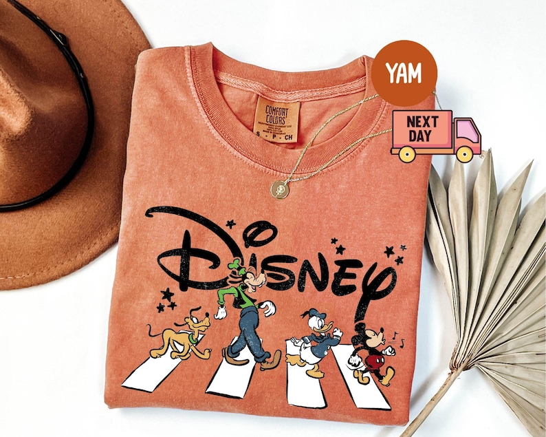 K&ouml;nnte beinhalten: Korallfarbenes T-Shirt mit dem Wort "Disney" in Schwarz, mit Zeichentrickfiguren, die einen Zebrastreifen &uuml;berqueren. Zu den Figuren geh&ouml;ren Pluto, Goofy, Donald Duck und Mickey Mouse. Eine goldene Halskette und ein brauner Hut sind ebenfalls sichtbar.