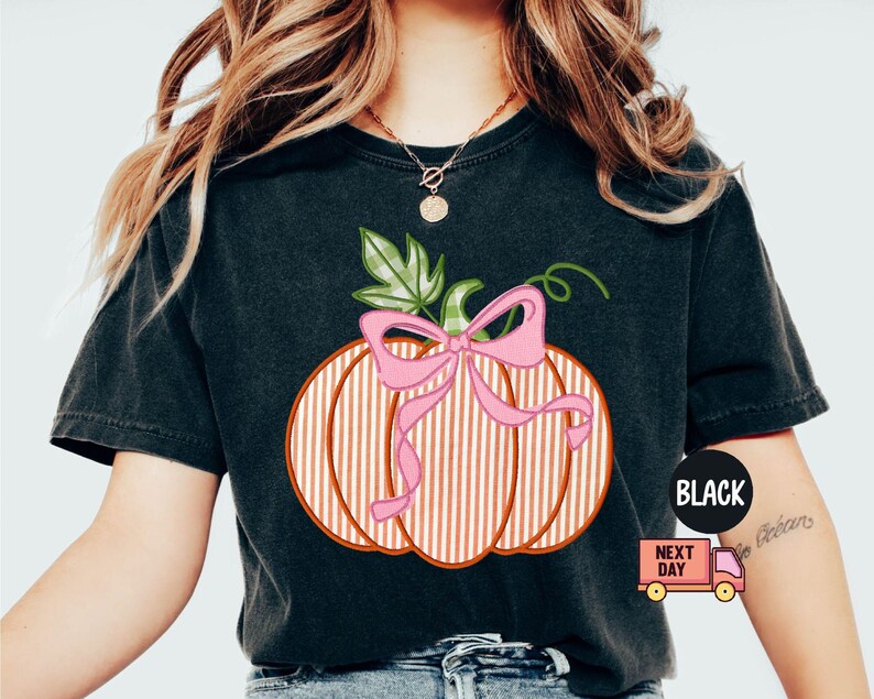 May include: Black t-shirt with a pumpkin design. The pumpkin has a pink bow and vertical stripes. A green leaf and stem are also present. The shirt has a round neck and short sleeves. The text "BLACK" is in a black circle.