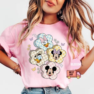 May include: Pink t-shirt featuring a floral design with cartoon characters. The flowers have checkered patterns and feature Daisy Duck, Donald Duck, Minnie Mouse, and Mickey Mouse. Butterflies and small flowers are also included in the design.