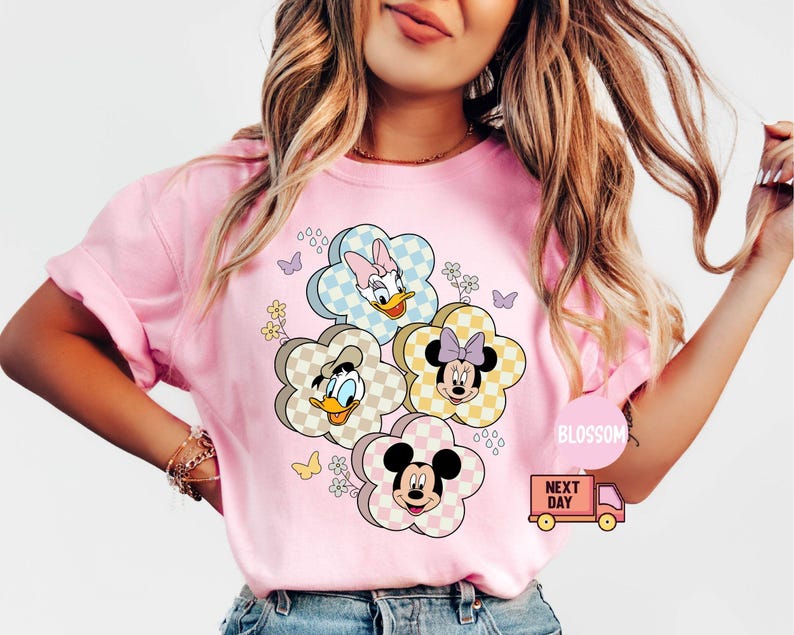 May include: Pink t-shirt featuring a floral design with cartoon characters. The flowers have checkered patterns and feature Daisy Duck, Donald Duck, Minnie Mouse, and Mickey Mouse. Butterflies and small flowers are also included in the design.
