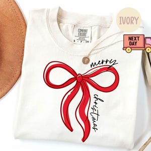 Può includere: T-shirt color avorio girocollo con un grande fiocco rosso e la scritta "merry Christmas" in nero. La maglietta ha una vestibilità ampia ed è realizzata in un materiale morbido. È visibile una collana d'oro.