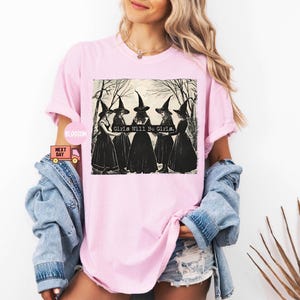 May include: Pink t-shirt with a black and white illustration of four witches wearing black pointed hats and robes. The text "Girls Will Be Girls." is printed on the shirt.