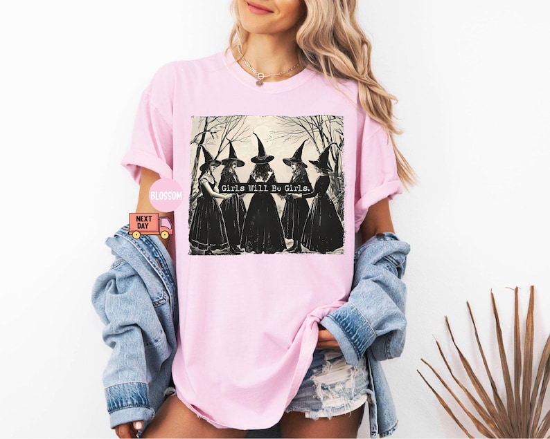 May include: Pink t-shirt with a black and white illustration of four witches wearing black pointed hats and robes. The text "Girls Will Be Girls." is printed on the shirt.