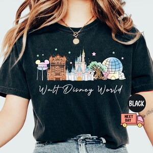 May include: Black t-shirt featuring a colorful graphic of Walt Disney World landmarks, including Cinderella Castle and Spaceship Earth. The words "Walt Disney World" are written in a script font below the graphic. The shirt is a dark color.