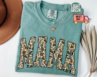 May include: Light green t-shirt with the word "MAMA" in a camouflage pattern. The shirt has a crew neck and short sleeves. A gold necklace with a small pendant is visible. The shirt is labeled "Comfort Colors".