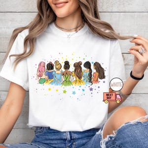 Puede incluir: Camiseta blanca con una ilustraci&oacute;n colorida de siete princesas de pie juntas. Las princesas llevan vestidos de diferentes colores y tienen diferentes peinados. La ilustraci&oacute;n est&aacute; rodeada de estrellas.