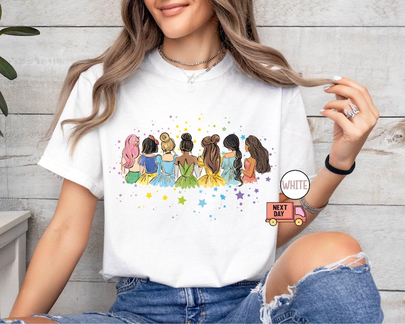 Puede incluir: Camiseta blanca con una ilustraci&oacute;n colorida de siete princesas de pie juntas. Las princesas llevan vestidos de diferentes colores y tienen diferentes peinados. La ilustraci&oacute;n est&aacute; rodeada de estrellas.