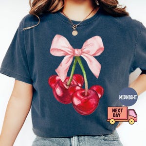Puede incluir: Camiseta azul marino oscuro con una ilustraci&oacute;n de acuarela de cerezas y un lazo rosa. Las cerezas son rojas y el lazo es rosa claro. La camiseta tiene un corte holgado.