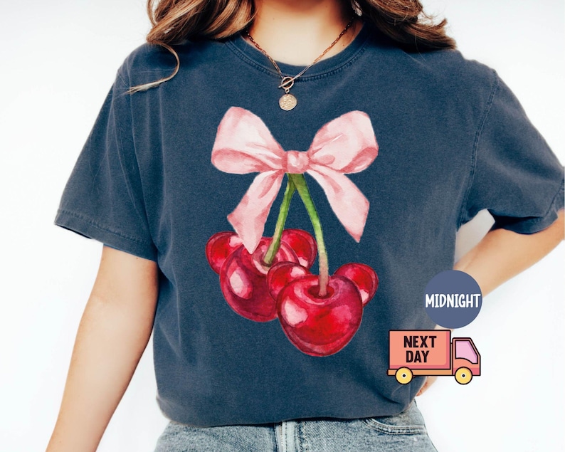 Puede incluir: Camiseta azul marino oscuro con una ilustraci&oacute;n de acuarela de cerezas y un lazo rosa. Las cerezas son rojas y el lazo es rosa claro. La camiseta tiene un corte holgado.