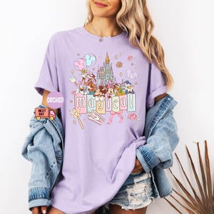 May include: A light purple t-shirt featuring a colorful graphic of a castle, cartoon characters, and the word "magical" in pastel letters. The design includes stars, balloons, and a magic wand. The shirt is worn by a person.