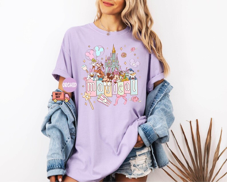 May include: A light purple t-shirt featuring a colorful graphic of a castle, cartoon characters, and the word "magical" in pastel letters. The design includes stars, balloons, and a magic wand. The shirt is worn by a person.