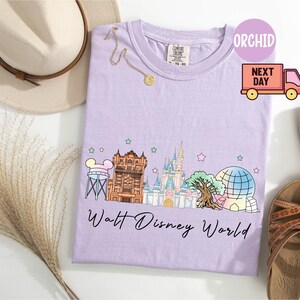 May include: A light purple t-shirt featuring a colorful graphic of Walt Disney World landmarks, including Cinderella's castle and Spaceship Earth. The text "Walt Disney World" is printed below the graphic. A gold necklace and a beige hat are also visible.