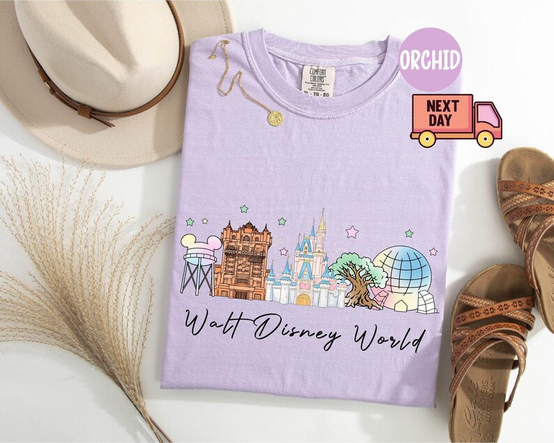 May include: A light purple t-shirt featuring a colorful graphic of Walt Disney World landmarks, including Cinderella's castle and Spaceship Earth. The text "Walt Disney World" is printed below the graphic. A gold necklace and a beige hat are also visible.