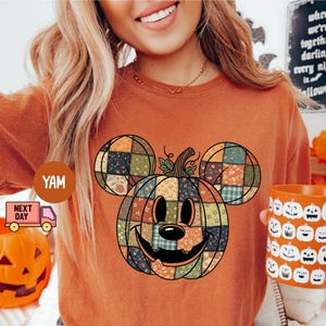 May include: Orange t-shirt featuring a patchwork pumpkin design with Mickey Mouse ears. The pumpkin has a smiling face and is surrounded by fall leaves. A matching orange mug with pumpkin faces is held in the hand.