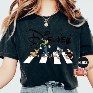 K&ouml;nnte beinhalten: Schwarzes T-Shirt mit Disney-Figuren, die eine Stra&szlig;e &uuml;berqueren, mit dem Disney-Logo dar&uuml;ber. Die Figuren sind Goofy, Pluto, Donald Duck und Mickey Mouse. Das T-Shirt hat einen Used-Look.