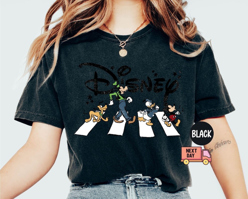 K&ouml;nnte beinhalten: Schwarzes T-Shirt mit Disney-Figuren, die eine Stra&szlig;e &uuml;berqueren, mit dem Disney-Logo dar&uuml;ber. Die Figuren sind Goofy, Pluto, Donald Duck und Mickey Mouse. Das T-Shirt hat einen Used-Look.