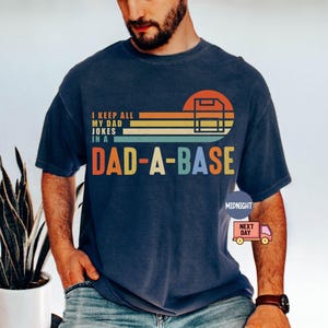 Könnte beinhalten: Marineblaues T-Shirt mit dem Text "I KEEP ALL MY DAD JOKES IN A DAD-A-BASE" in einem Retro-Design. Die Grafik enthält einen Sonnenuntergang und eine Computer-Server-Illustration. Ein bequemes, lässiges Kleidungsstück.