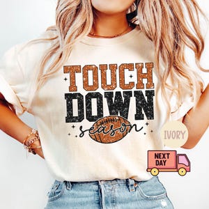 May include: A white t-shirt with the text "Touch Down Season" in black and brown glitter lettering. A brown and white football graphic is centered below the text. The shirt is available in ivory.