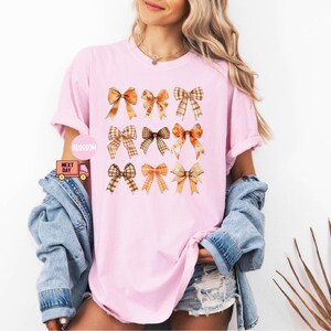 May include: A pink t-shirt with a pattern of nine orange and brown bows. The bows are arranged in three rows of three.