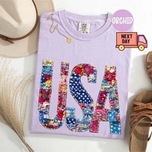 May include: A light purple t-shirt with the word "USA" in a patchwork design featuring floral and star-spangled patterns. The shirt is accompanied by a straw hat, sandals, and a delicate gold necklace. The image also includes a "Next Day" delivery graphic.