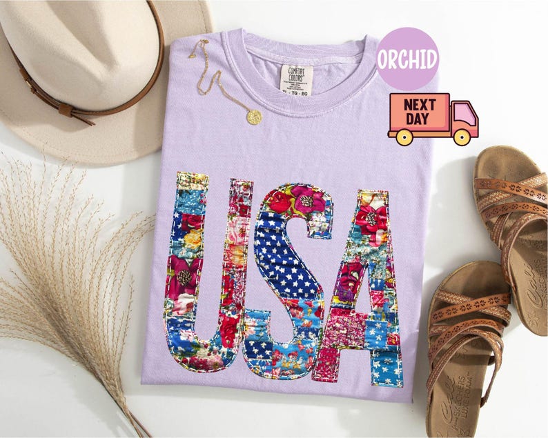 May include: A light purple t-shirt with the word "USA" in a patchwork design featuring floral and star-spangled patterns. The shirt is accompanied by a straw hat, sandals, and a delicate gold necklace. The image also includes a "Next Day" delivery graphic.