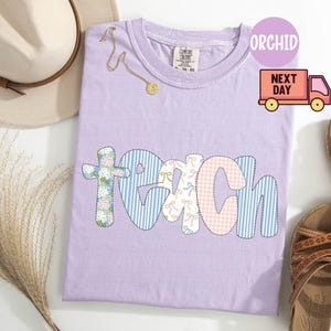 May include: A light purple Comfort Colors t-shirt with the word "TEACH" in a patchwork design. The letters are in various patterns, including floral, blue stripes, and pink gingham. A gold necklace and a beige hat are also visible.