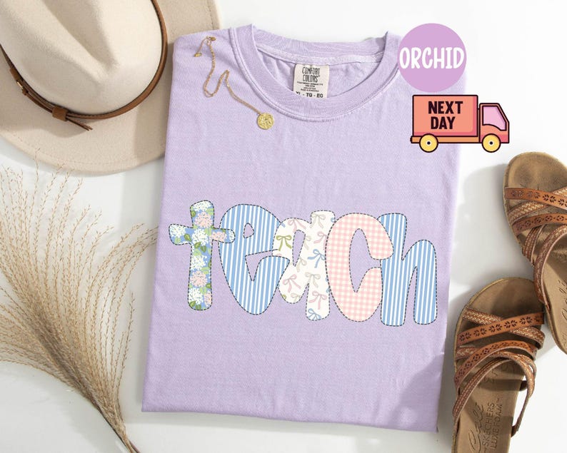 May include: A light purple Comfort Colors t-shirt with the word "TEACH" in a patchwork design. The letters are in various patterns, including floral, blue stripes, and pink gingham. A gold necklace and a beige hat are also visible.