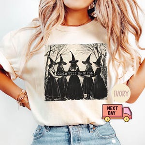 May include: A white t-shirt with a black and white illustration of four witches wearing black dresses and pointy hats. The text "Girls Will Be Girls." is printed on the shirt.