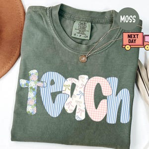 May include: Moss green Comfort Colors t-shirt with the word "TEACH" appliqued in floral, striped, and gingham patterns. A gold necklace with a pendant rests on the shirt. The shirt is folded, with a brown hat and dried plant in the background.