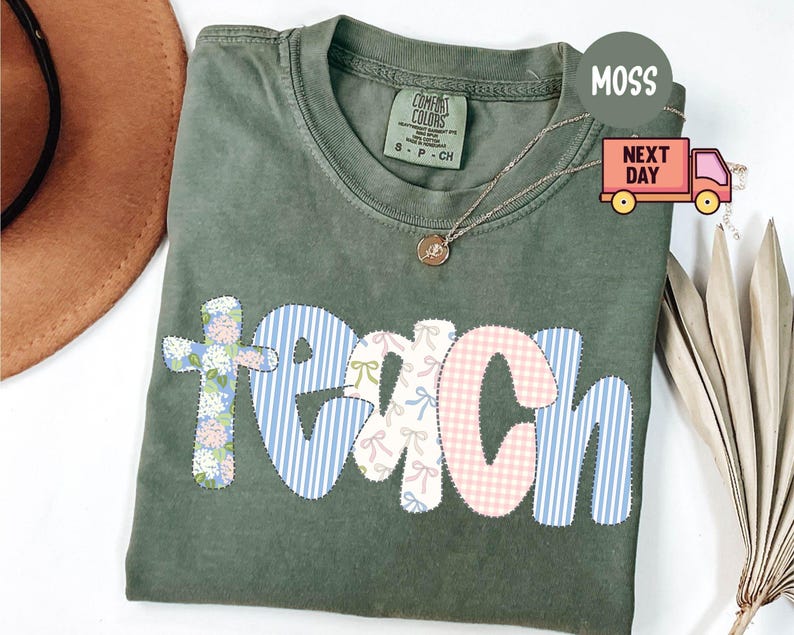 May include: Moss green Comfort Colors t-shirt with the word "TEACH" appliqued in floral, striped, and gingham patterns. A gold necklace with a pendant rests on the shirt. The shirt is folded, with a brown hat and dried plant in the background.