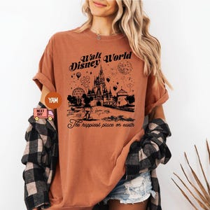 May include: A rust-colored t-shirt featuring a black graphic of Cinderella's castle, Mickey Mouse, and the text "Walt Disney World" and "The happiest place on earth". The shirt has short sleeves and a relaxed fit.