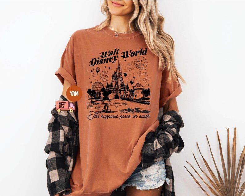 May include: A rust-colored t-shirt featuring a black graphic of Cinderella's castle, Mickey Mouse, and the text "Walt Disney World" and "The happiest place on earth". The shirt has short sleeves and a relaxed fit.