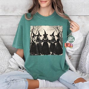 May include: A teal green t-shirt with a vintage illustration of four witches wearing black dresses and pointed hats. The text "Girls Will Be Girls." is printed below the witches.