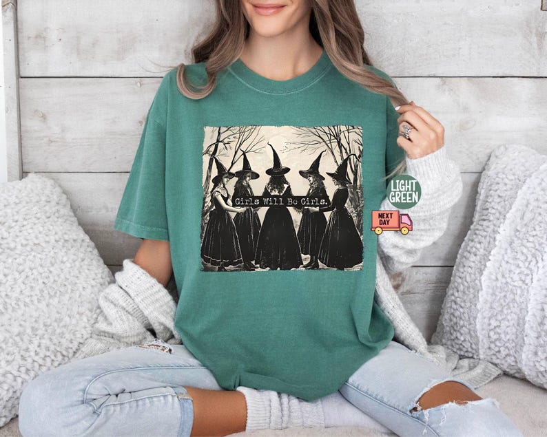May include: A teal green t-shirt with a vintage illustration of four witches wearing black dresses and pointed hats. The text "Girls Will Be Girls." is printed below the witches.