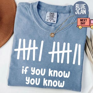 May include: A blue jean colored t-shirt with white text that reads "if you know you know". The shirt features a tally mark design. A gold necklace is draped on the shirt. The shirt is a Comfort Colors brand.